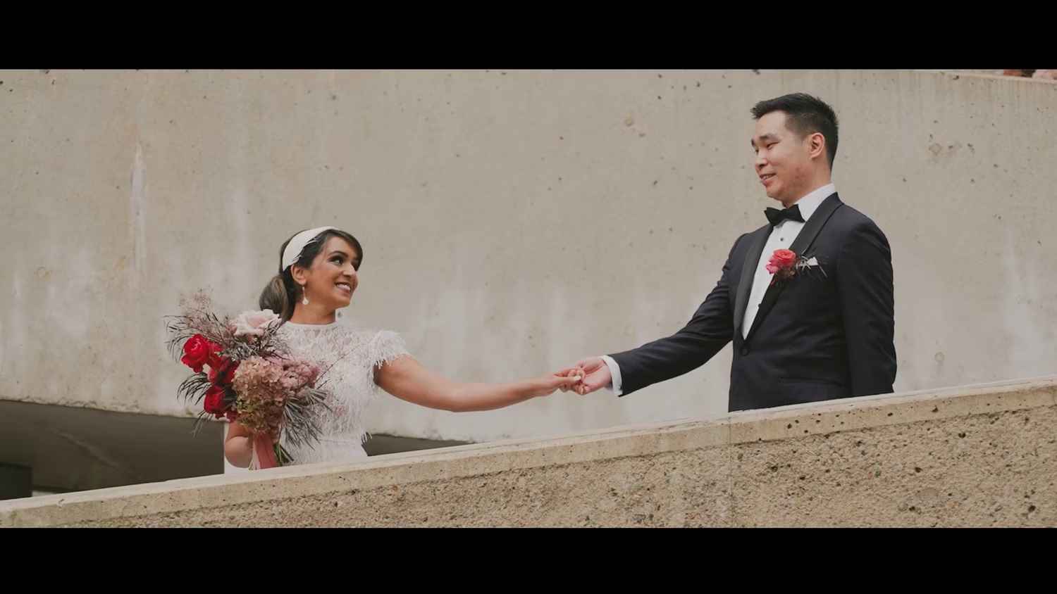 Bride and groom holding hands on a balcony, bride carrying a red bouquet